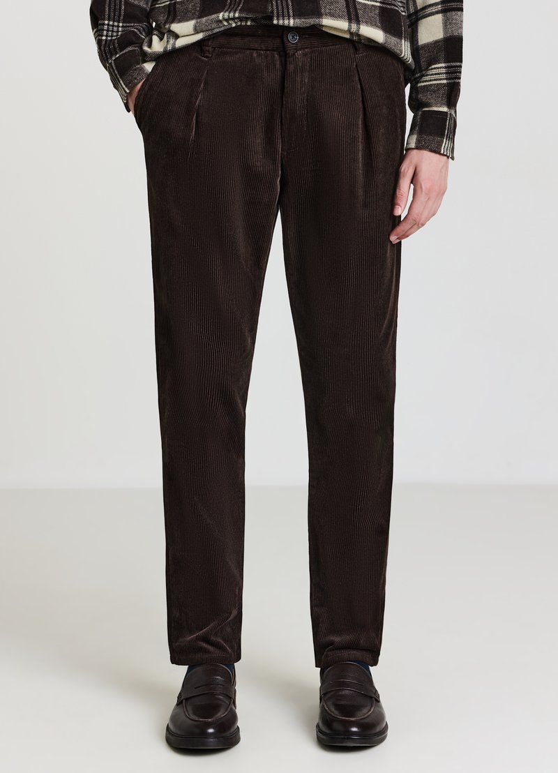 Homme portant un pantalon en velours côtelé brun foncé, des mocassins noirs et une chemise à carreaux noir et blanc, les mains dans les poches.