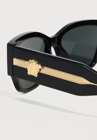Μαύρα γυαλιά ηλίου με χοντρά πλαίσια, σκουρόχρωμους φακούς και χρυσή διακοσμητική λεπτομέρεια με κεφάλι Μέδουσας στα πλαϊνά.