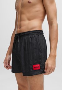 Schwarze Schwimmshorts aus leichtem Stoff, mit elastischem Bund, Kordelzug und einem roten Logo-Patch.