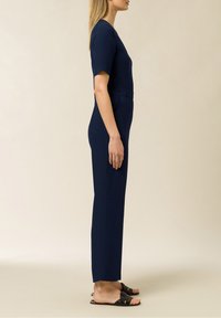 Tuta navy con maniche corte, scollatura a V e silhouette aderente. Tessuto morbido e gambe dritte, abbinata a sandali neri con fascia.