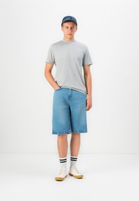 Giovane uomo con una maglietta grigia, pantaloni corti di jeans, calze a righe, scarpe da ginnastica e un cappellino blu, in piedi su uno sfondo bianco.