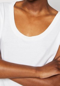 Valkoinen lyhythihainen t-paita, jossa on leveä, pyöreä pääntie ja pehmeä, sileä rakenne. Mallin kädet ristissä, paljastaen ihonsävyn.