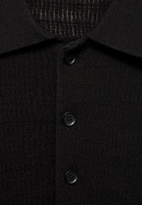 Polo shirt nera textured con colletto, tre bottoni neri e un tessuto a fantasia contrastante nell'area del collo.