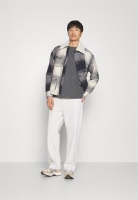 Geruit jasje met een crèmekleurige neplederen kraag, grijze t-shirt eronder, witte broek en beige sneakers met grijze accenten.