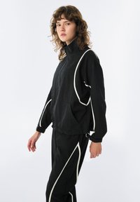 Giacca nera con zip e rifiniture bianche, caratterizzata da una vestibilità comoda, grande tasca frontale e maniche lunghe. Il tessuto sembra avere una texture.