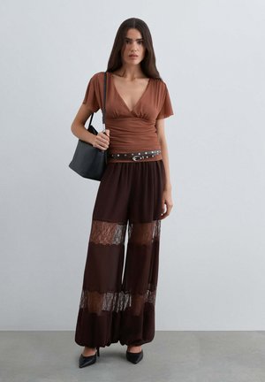 Mujer con top de escote en V color marrón óxido, pantalones anchos con paneles de encaje, cinturón negro con tachuelas, tacones negros y bolso negro de hombro.