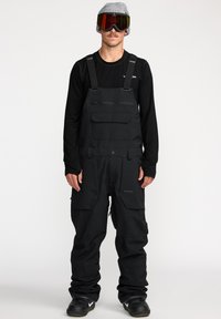 Zwarte sneeuw_overalls met verstelbare banden, ritszakken en een relaxte pasvorm. Gepaard met een zwart langemouwen shirt en een skibril.