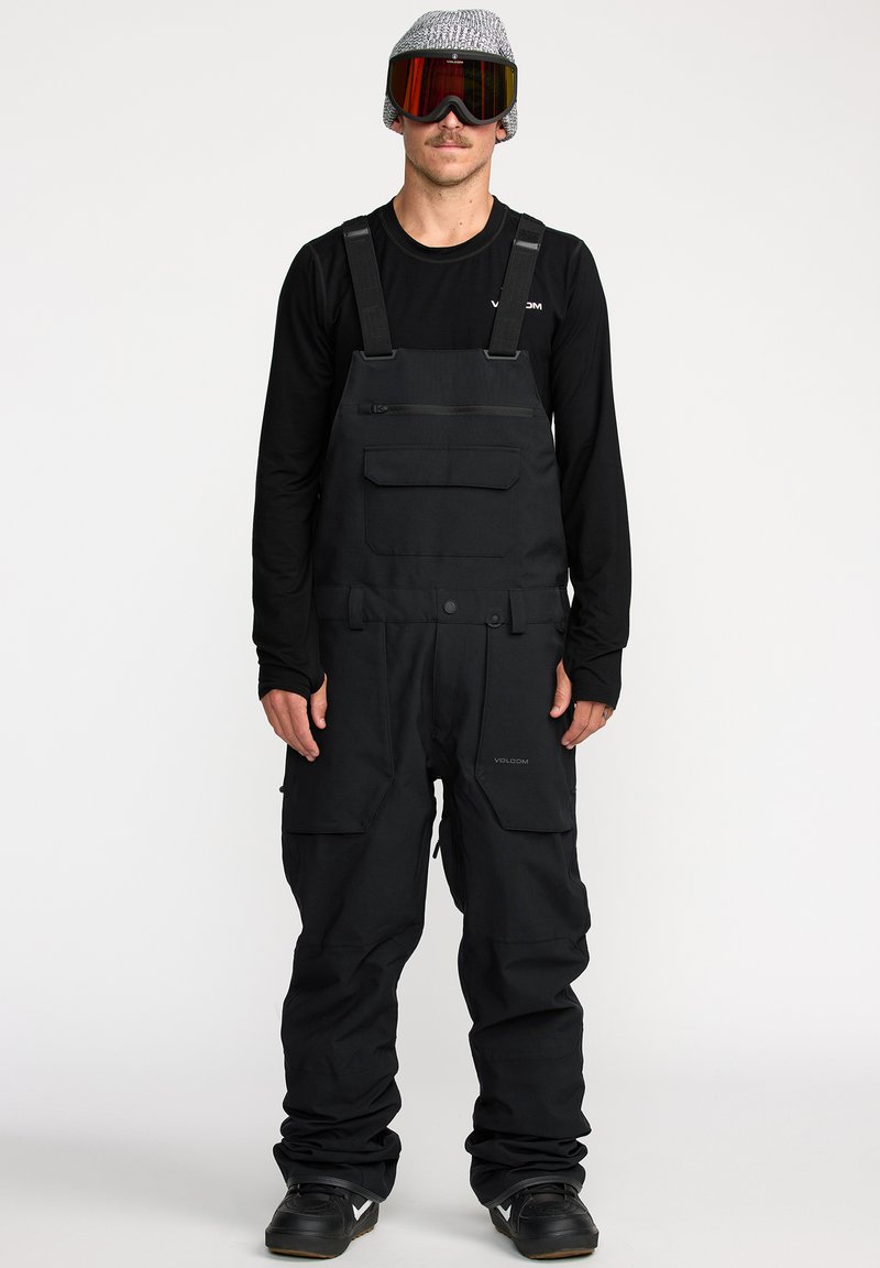 Zwarte sneeuw_overalls met verstelbare banden, ritszakken en een relaxte pasvorm. Gepaard met een zwart langemouwen shirt en een skibril.