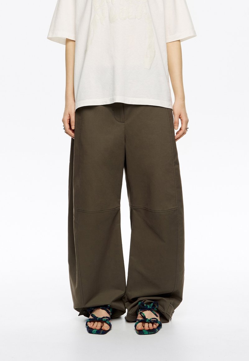 Pantalones de pierna ancha color verde oliva con una textura suave y dos costuras visibles. Combinados con sandalias de diseño estampado con correas anudadas.