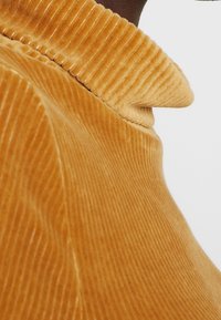Sennepsgul corduroy-stof med lodrette ribber, der har et højt halsdesign og en blød tekstur. Fokuser på stofdetaljer og farve.