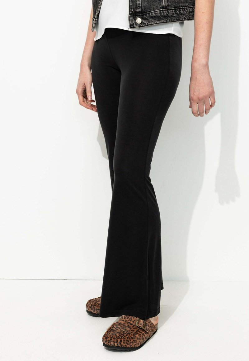 Leggings noires évasées en tissu extensible, présentant une texture lisse. Associés à des sabots à imprimé léopard marron pour un contraste supplémentaire.