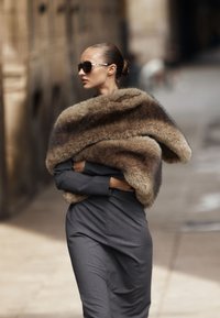 Donna con occhiali da sole scuri e stole di pelliccia sopra un vestito grigio, che cammina all'aperto con le braccia incrociate, guardando di lato.