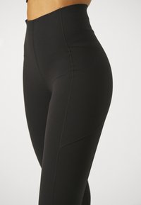 Høyt liv svarte leggings laget av elastisk stoff med flatlocksømmer og en glatt tekstur, med en tettsittende silhuett.