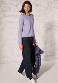 Hellblau, gerippter Langarm-Oberteil kombiniert mit dunkelblauen, geraden Jeans. Weiße Sneakers mit grünen Akzenten, hält eine lila Pufferjacke.