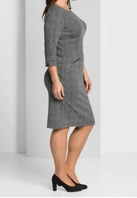 Robe à carreaux gris avec une silhouette ajustée, des manches trois-quarts et une fermeture éclair latérale. Portée avec des chaussures à talons noirs.