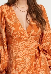 Oranje wikkeljurk met een bloemenpatroon, voorzien van lange mouwen, een diepe V-hals en een strikdetail bij de taille. Zacht, textuurachtig materiaal.