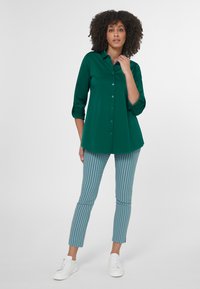 Groene overhemdd met opgerolde mouwen, gecombineerd met gestreepte blauw-witte broek tot de enkel en witte sneakers.