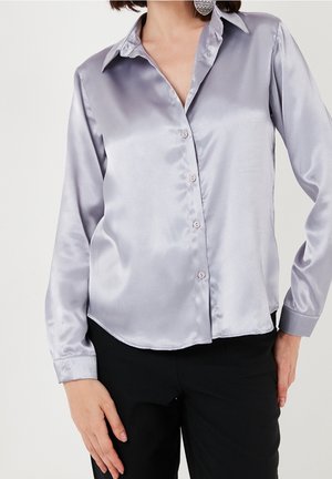 Femme portant une chemise en satin argenté à manches longues boutonnée et un pantalon noir, debout avec une main le long du corps.