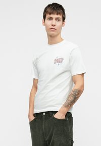 Mladý muž s krátkymi hnedými vlasmi, oblečený v bielom tričku "Santa Cruz" a tmavozelených manšestrových nohaviciach, s rukami v vreckách.