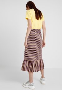 Gemusterter Midirock mit schwingendem Saum, der ein geometrisches Muster in Lila, Gelb und Pink zeigt. Kombiniert mit einem kurzärmeligen gelben Oberteil und Sneakers.