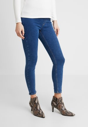 Femme portant un jean skinny bleu et des bottines en cuir effet peau de serpent marron, debout devant un fond clair uni.