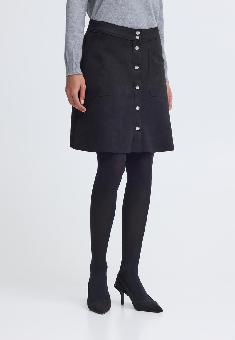 Frau trägt einen schwarzen Rock mit durchgehendem Knopfverschluss und Taschen, schwarze Strumpfhosen, schwarze High Heels und einen hellgrauen Pullover mit langen Ärmeln vor einem schlichten Hintergrund.