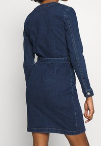 Jeansklänning, mörkblå, lång ärm, åtsittande midja med bälte, enkel krage och sömnadsdetaljer längs sömmarna.