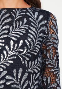 Mørkeblå kjole med intrikate paljettblomsterbroderier på blonde, lange, gjennomsiktige ermer og rund halsåpning.