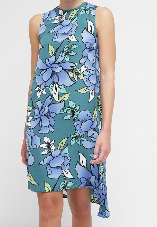 Robe sans manches couleur sarcelle avec un grand imprimé floral bleu et blanc et un ourlet asymétrique portée par une personne avec une main détendue le long du corps.