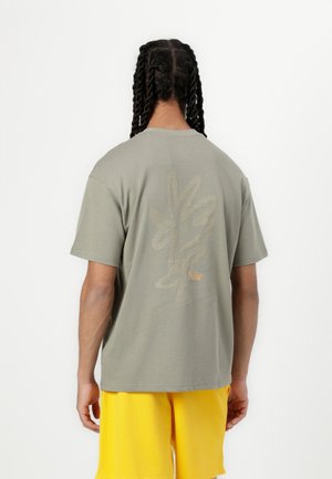 Personne avec des cheveux tressés, portant un t-shirt gris Nike avec un motif de feuille et un short jaune, vue de dos sur un fond uni.