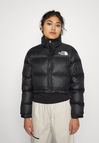 Jachetă puffer neagră, scurtă, cu guler înalt, având fermoar frontal și accent de logo. Textură netedă, lucioasă, cu model matlasat.