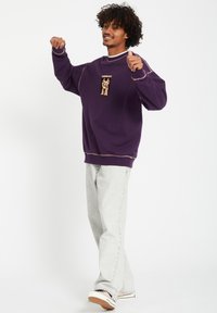 Paarse sweatshirt met een grafische print aan de voorkant, contrasterende stiksels en ribgebreide manchetten. Draag het met lichtgrijze jeans en sneakers, staand in een ontspannen houding.