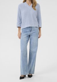 Hellblauer, gestrickter V-Ausschnitt-Pullover mit Dreiviertelärmeln, kombiniert mit hochtaillierten, weiten, hellen Denim-Jeans und schwarzen, spitzen Schuhen.