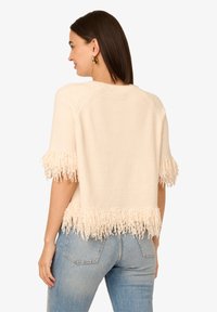 Cremfarbener Pullover mit Fransenverzierungen an den Ärmeln und am Saum. Strukturierter Stoff und boxy Schnitt. Getragen mit Jeans, die Rückansicht wird gezeigt.