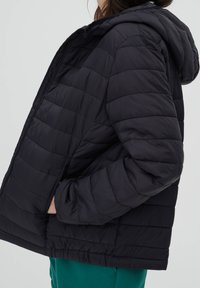 Personne portant une veste matelassée noire à capuche, les mains dans les poches latérales, par-dessus un pantalon vert, vue de profil sur un fond uni.