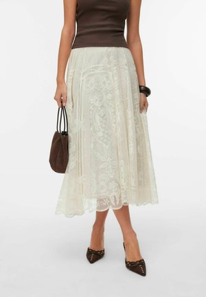 Femme portant une jupe midi en dentelle crème, un top marron sans manches, des talons pointus marron, tenant un sac à main marron avec des poignées noires.