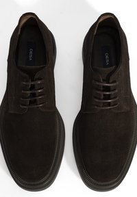 Une paire de chaussures de ville pour hommes en suède marron foncé, à lacets, avec des semelles en caoutchouc noir, vues de dessus sur une surface blanche.