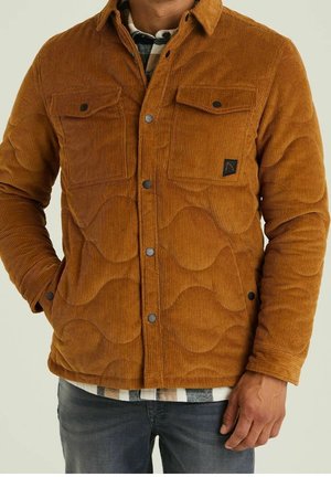 Mann trägt eine braune Cord-Steppjacke mit Druckknöpfen, zwei Brusttaschen, darunter ein kariertes Hemd und blaue Jeans.