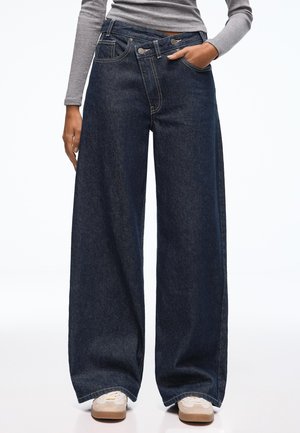 Wide-leg dark denim jeans with a high waist, featuring prominent stitching, two front pockets, and a button closure design.