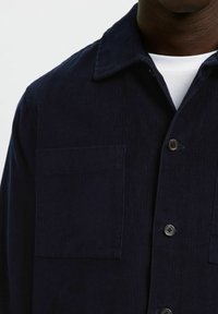 Chaqueta de pana azul oscuro con un bolsillo en el pecho y botones negros, llevada sobre una camiseta blanca de cuello redondo.