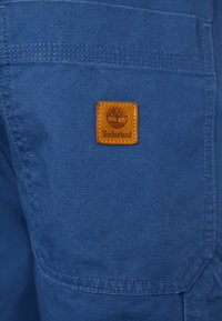 Niebieskie szorty z tkaniny z przeszywaną kieszenią, z brązową skórzaną naszywką Timberland. Gładka tekstura i swobodne detale projektu.