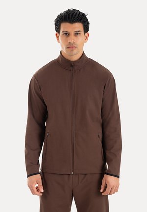 Mann mit dunklen Haaren, trägt eine braune Jacke mit Reißverschluss und passende Hose, steht vor einem schlichten hellen Hintergrund.