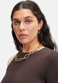 Guldsmeds kæde med store led, båret af en model med mørkt hår, iført en brun ribbet top. Enkle hoopearrrings.