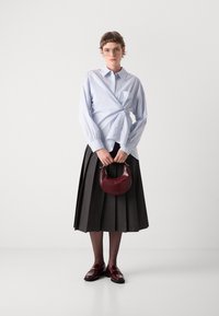 Chemise bleu clair nouée à la taille, jupe plissée noire, sac à main bordeaux avec motif crocodile et mocassins assortis bordeaux. Lunettes portées.