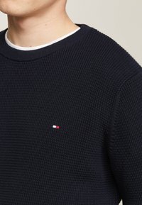 Maglione blu navy a maglia con texture a coste, scollo rotondo e piccolo logo rosso, bianco e blu sul petto; vestibilità classica e design minimale.