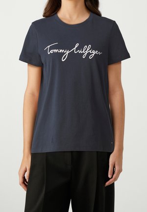 Femme portant un t-shirt bleu marine à manches courtes avec un logo "Tommy Hilfiger" en écriture cursive blanche et un pantalon noir, debout devant un fond uni.