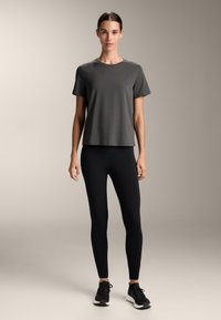 Temno siva kratka majica s kratkimi rokavi v kombinaciji z črnimi legicami in črnimi športnimi čevlji, postavljena na nevtralni ozadju.