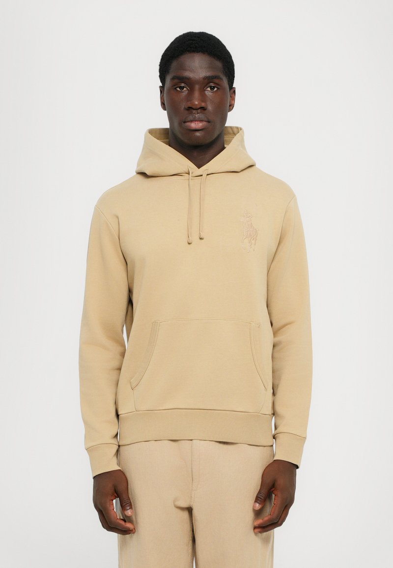 Beige hoodie tillverkad av mjukt tyg, med en framficka, dragsko och en broderad logotyp på vänster bröst. Texturen är slät.