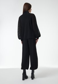 Zwarte trui met netmouwen, hoge hals, gecombineerd met zwarte wijde broek met netpaneeltjes aan de zijkanten. Draagt chunky platformlaarzen.