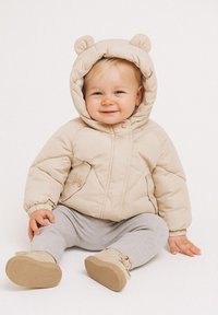 Beige padded jacket with rounded hood featuring bear ears, zip closure, diagonal stitching, worn over gray pants and shoes. Child sitting.